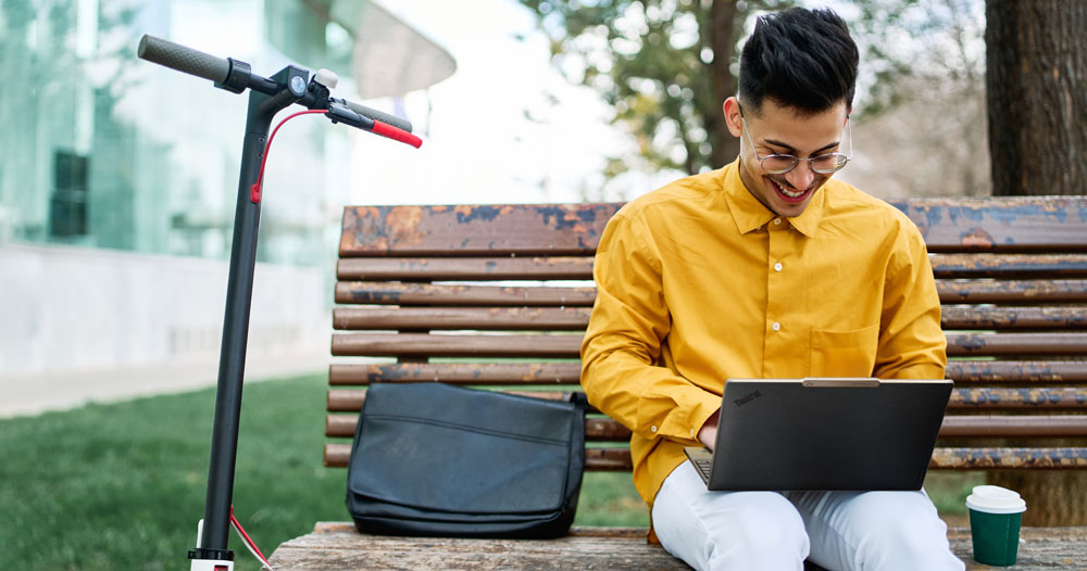 ein junger Mann in gelber Jacke sitzt auf einer Bank. Neben ihm steht ein Kaffeebecher. An einer Laterne lehnt ein E-Scooter
