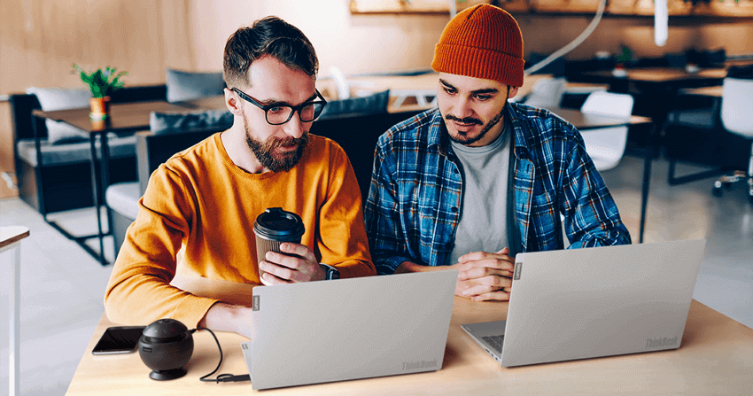 Zwei junge Männer sitzen mit Coffee-To-Go-Bechern vor einem Laptop. Einer scheitn dem anderen etwas zu zeigen